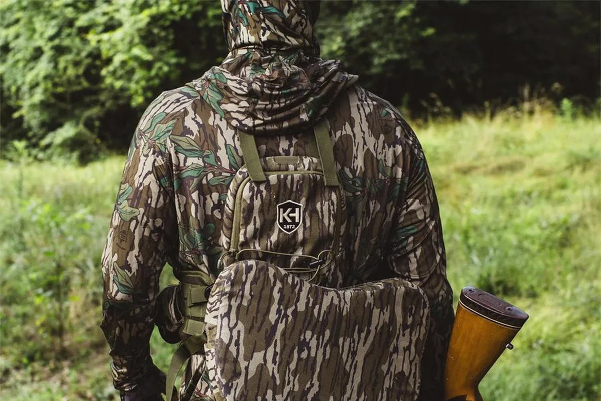 Hunter in full Mossy Oak Bottomland camo layering system walking a green field edge with a shotgun